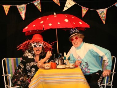 Angelika Dormeyer und Arwed Werner sitzen an einem Tisch auf der Bühne des Muh-Theaters in lustigen Kostümen zum Thema Camping.