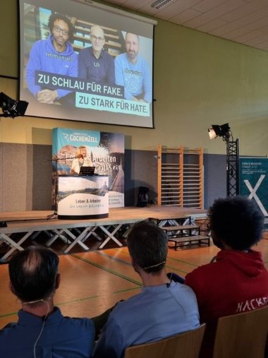 Cyber Event Landrätin Beilstein hält ihre Eröffnungsrede, man sieht die Hinterköpfe von v.l.n.r. Ulrich Irnich, Dr. Christian Reinhardt und Immanuel Bär, die auf die Bühen schauen.