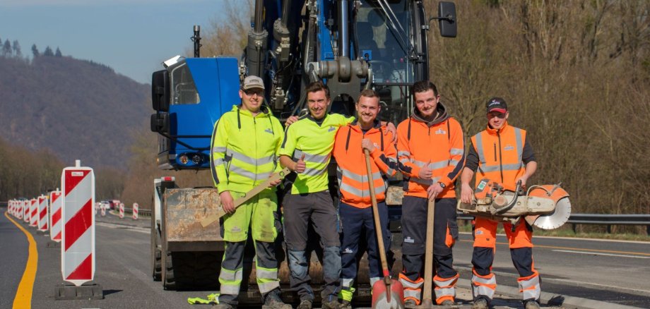 Fünf Azubis stehen mit Werkzeugen in der Hand vor einem Bagger von Schnorpfeil.