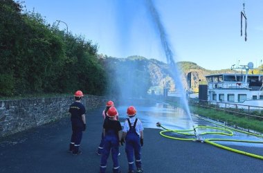 Drei Feuerwehrkinder bei einer Übung, eine Wasserfontäne schießt aus eienm Schlauch in die Luft.