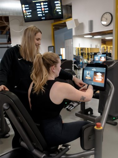 Trainieren im FusionFitness Eine Trainerin bei der Einweisung einer Kundin auf einem Fitnessgerät