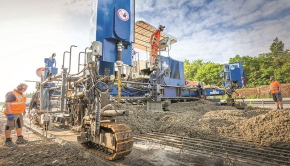 Fahrbahnbeton Bauarbeiten an der Autobahn, eine große Maschine sowie zwei Arbeiter sind zu sehen.