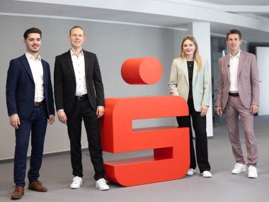Gruppenfoto des Sparkassenteams in Cochem Azubi Admir, Jan Eric Schütze, Azubine Mina und Frank Michels stehen am Sparkassenlogo